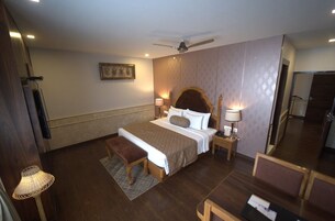 A hotel room with a large bed, a wooden bench, a desk with chairs, a ceiling fan, and a wall-mounted TV.