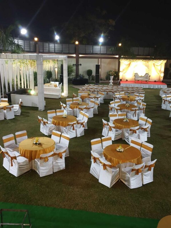 An outdoor event space with round tables and chairs, a stage with a backdrop, and a seating area with white chairs and gold sashes. - Hotel JSR Continental (Dehradun)