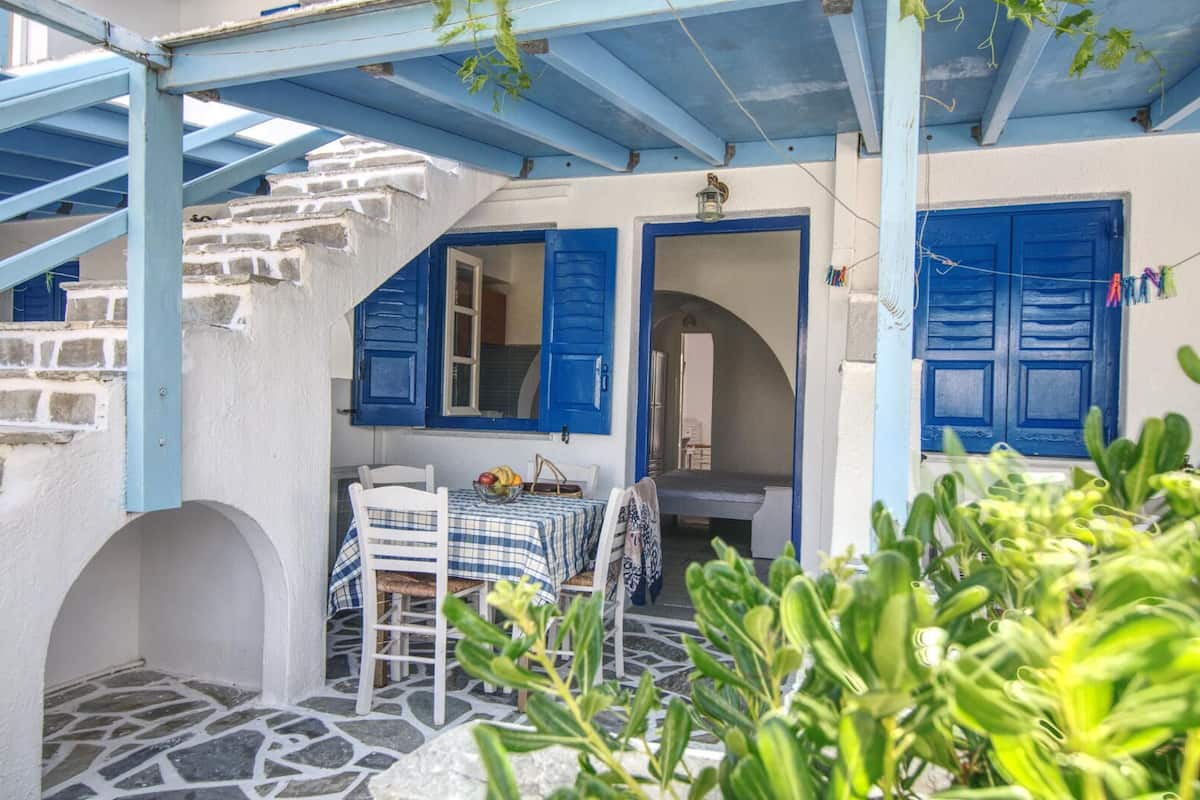 A covered outdoor seating area with a table and chairs, a stone floor, and blue shutters on the walls.