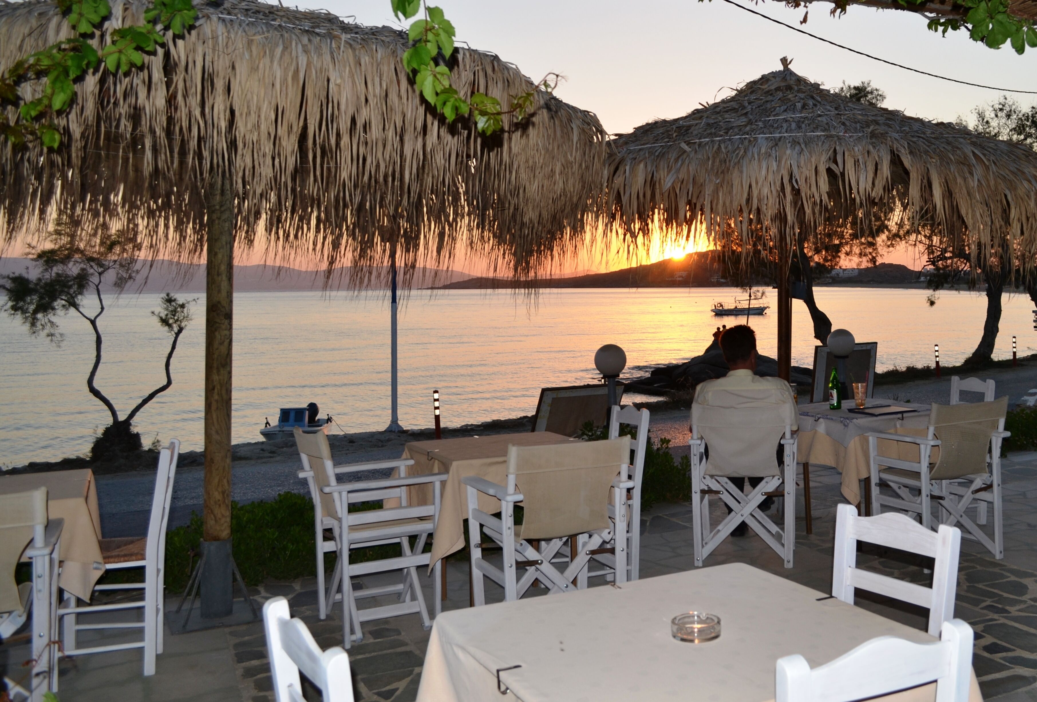 An outdoor dining area with tables and chairs, a thatched umbrella, and a view of the sunset over water.
