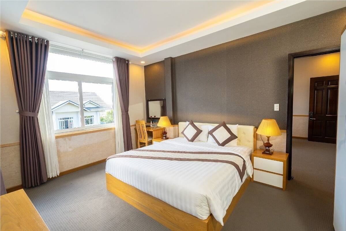 A hotel room with a large bed, two bedside tables with lamps, a window with curtains, and a view of buildings outside.