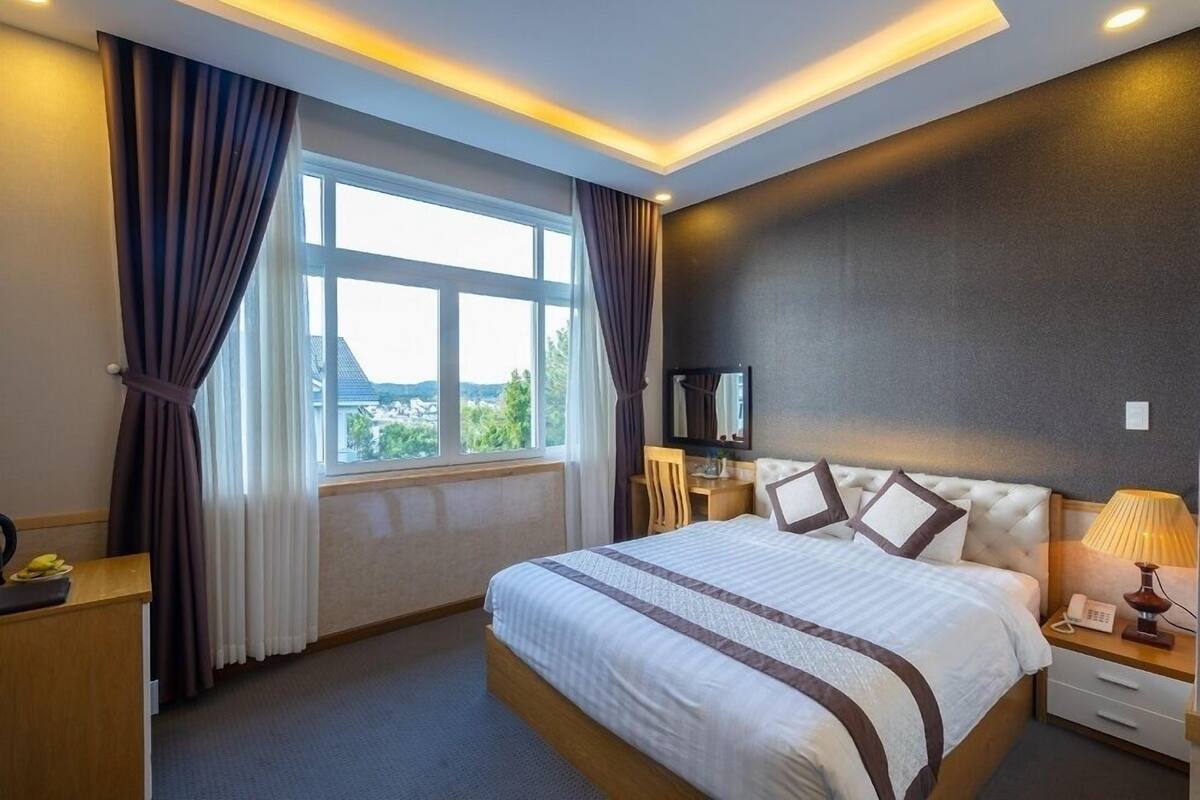 A hotel room with a large bed, a desk, a chair, a bedside table with a lamp, and a view of the outdoors through a window.