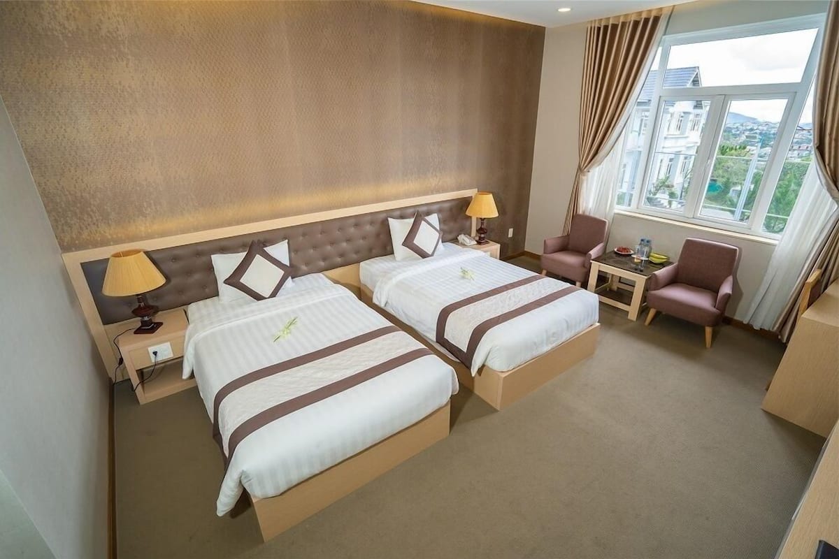 A hotel room with two beds, a small table, a chair, and a window with a view.