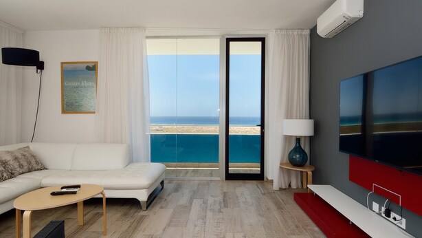 A modern hotel room with a white sofa, a small coffee table, a TV, and a large window offering a view of the beach. - Luxe Loft Casa Atlantica Morro Jable 658 BY PVL (Morro Jable)