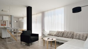 A modern living room with a white sofa, a black armchair, and a coffee table. There is a kitchen area with a dining table and chairs visible in the background. - Luxe Loft Casa Atlantica Morro Jable 658 BY PVL (Morro Jable)