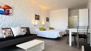 Een moderne hotelkamer met een gestructureerde witte muur, twee bedden met groen-witte beddengoed, een zwarte bank met bloemenkussens, een witte salontafel, en een eethoek met een stoel en tafel.