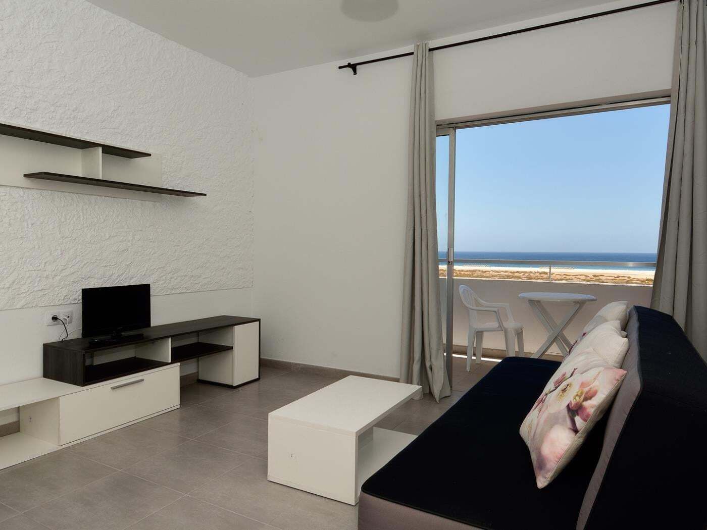 A modern room with a TV, a sofa, a dining table, and a balcony with a view of the beach.