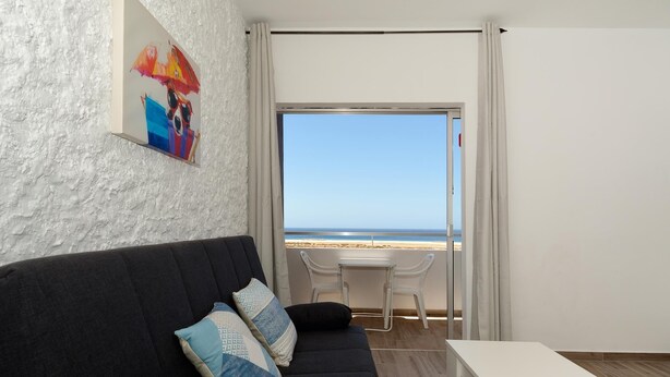 Une pièce avec un canapé sombre, une table blanche et une vue sur la plage, visible à travers une grande fenêtre.