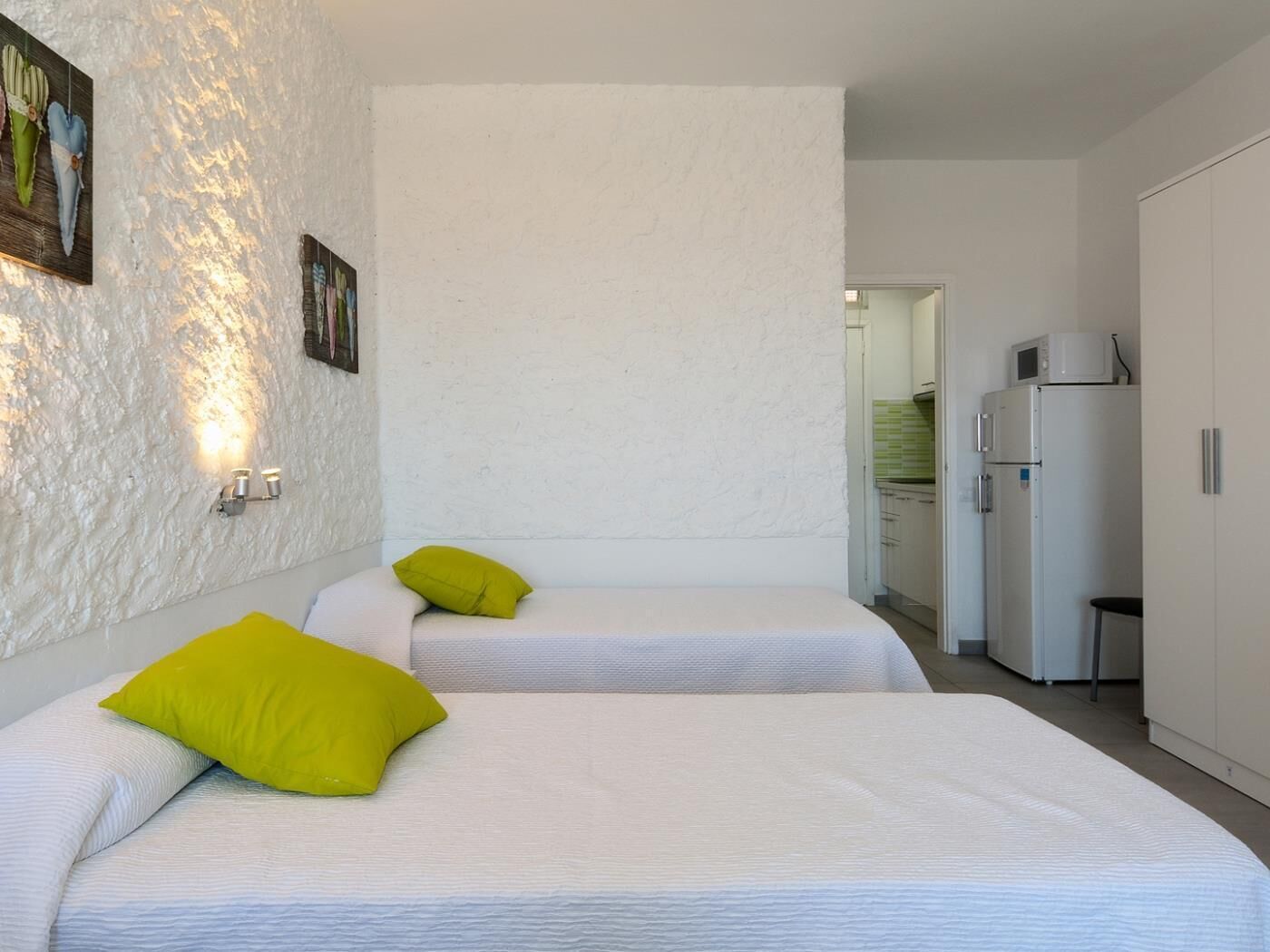 Un dormitorio moderno con dos camas, una pared blanca con textura, una pequeña kitchenette y una puerta que da a un baño.