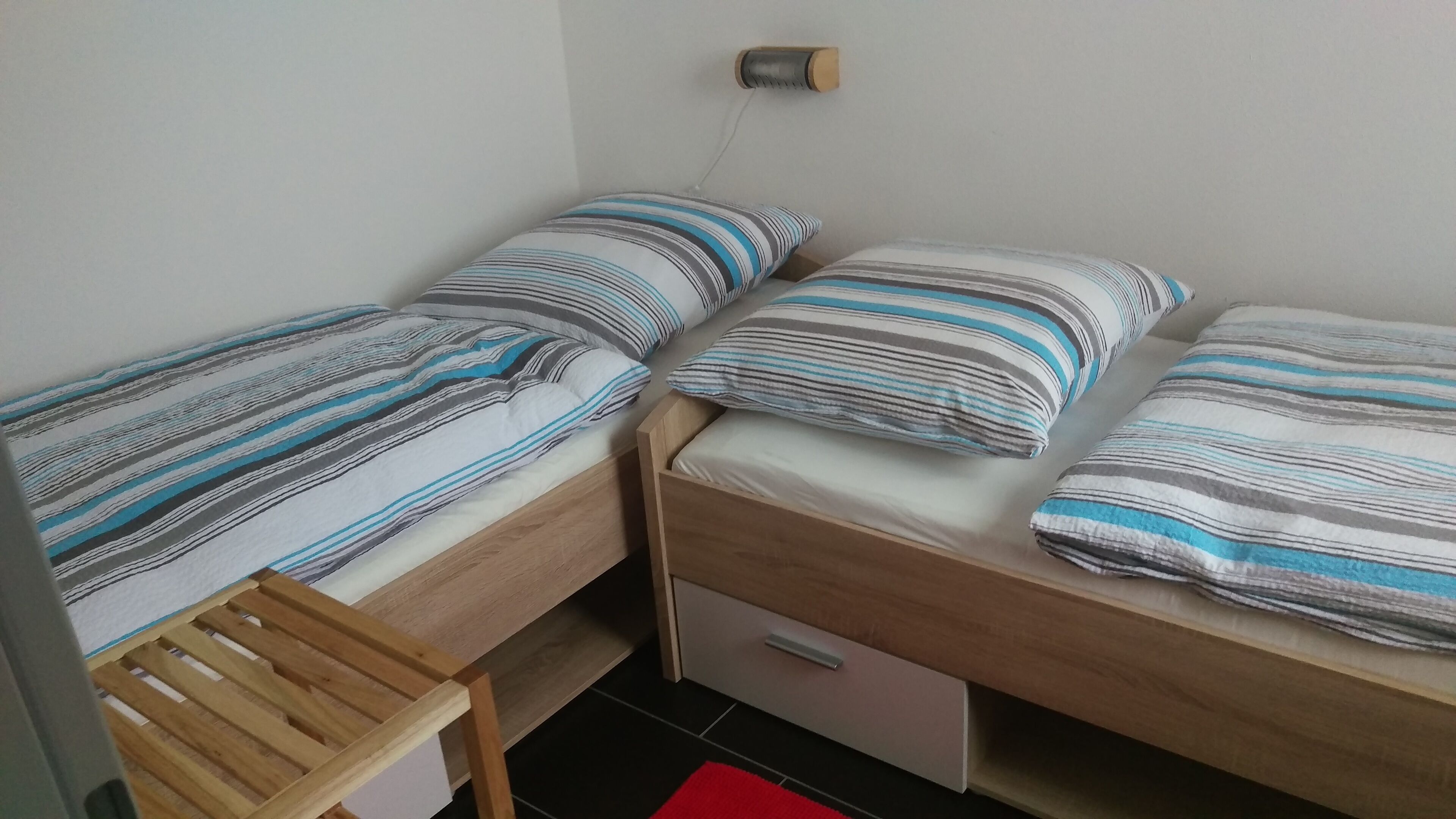 Ett litet, kompakt rum med två enkelsängar, båda med randiga sängkläder. Vid fotändan av en av sängarna står en träbänk, och under den andra sängen finns en liten byrå med lådor.