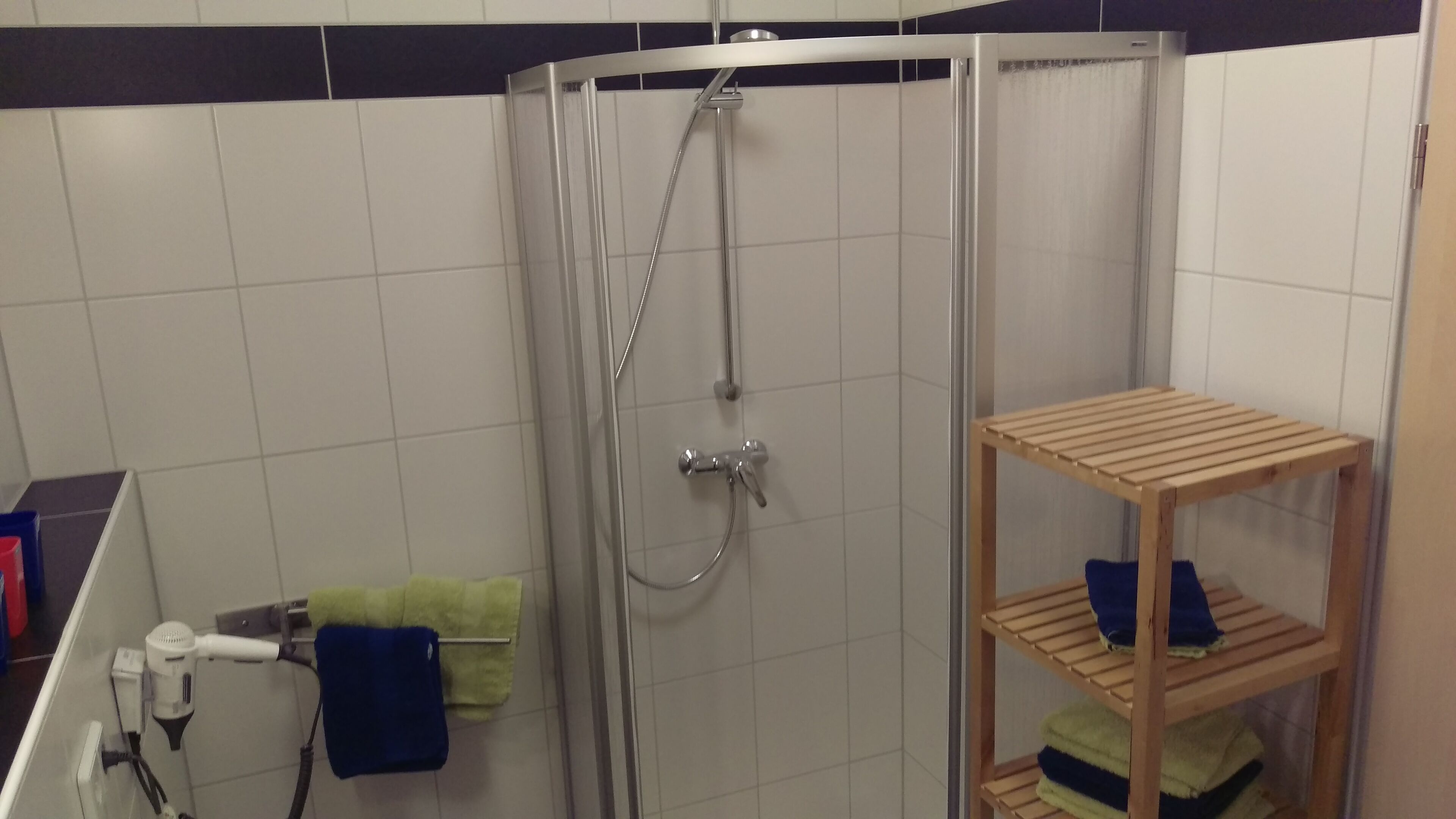 A shower area with a glass door, a wooden shelf with towels, and a hairdryer on a counter.
