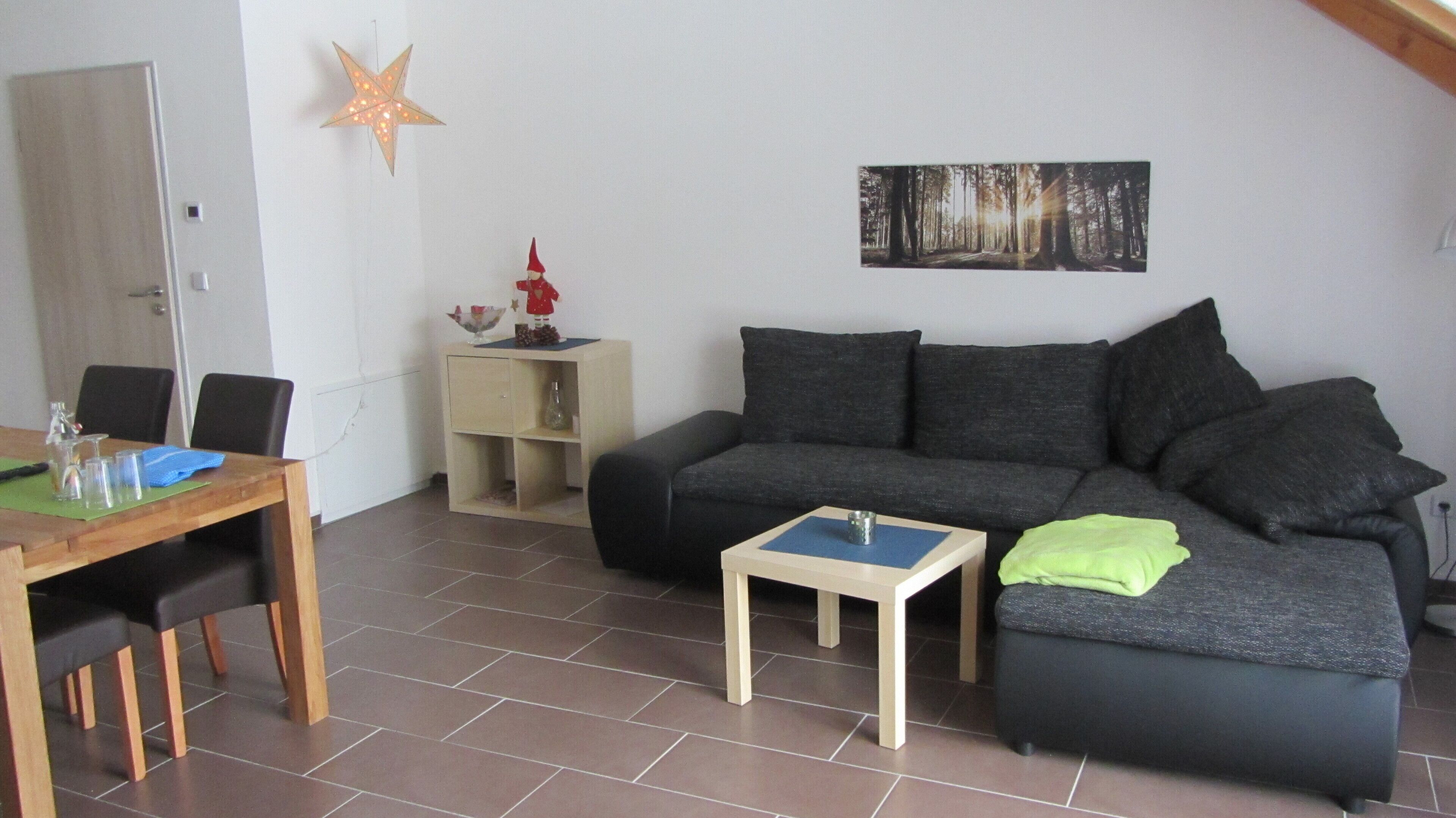 A living room with a dark grey sofa, a small wooden coffee table, a dining table with chairs, a star-shaped light fixture, and a forest-themed painting on the wall.