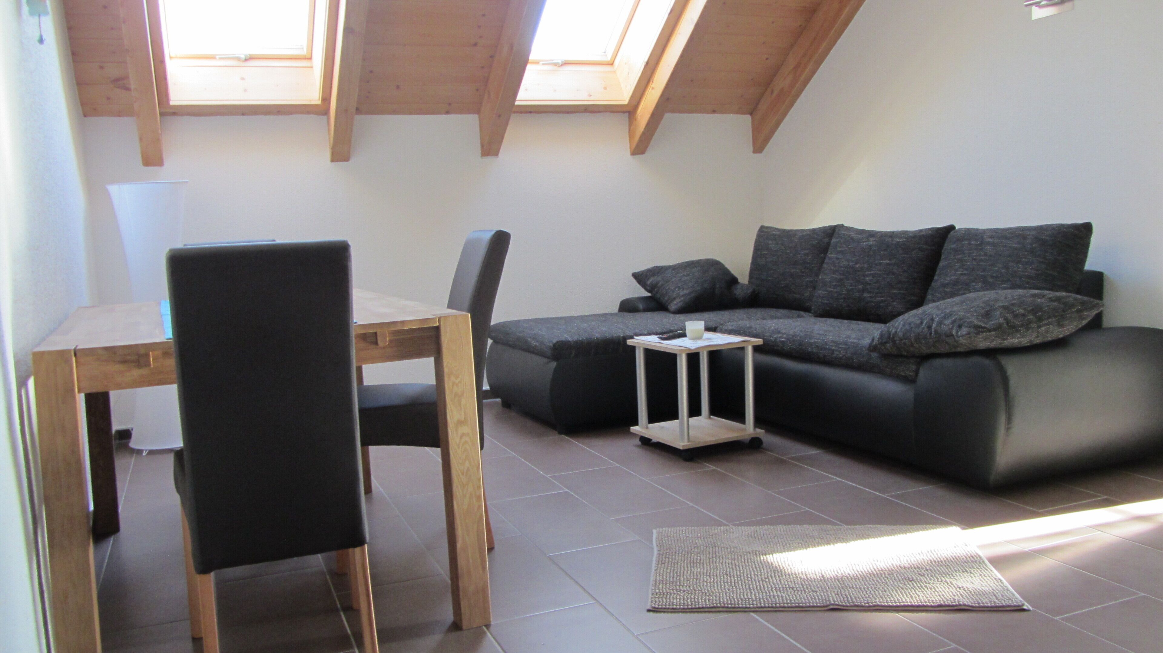 A small, modern living space with a wooden dining table, a black chair, a grey sofa with cushions, and a small white side table.