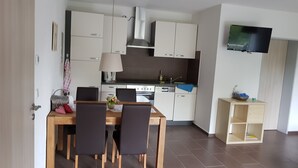 A modern kitchen with white cabinets, a stove, and a dishwasher. There is a dining table with chairs and a TV mounted on the wall. - Ferienwohnung Neuenbau (Föritztal)