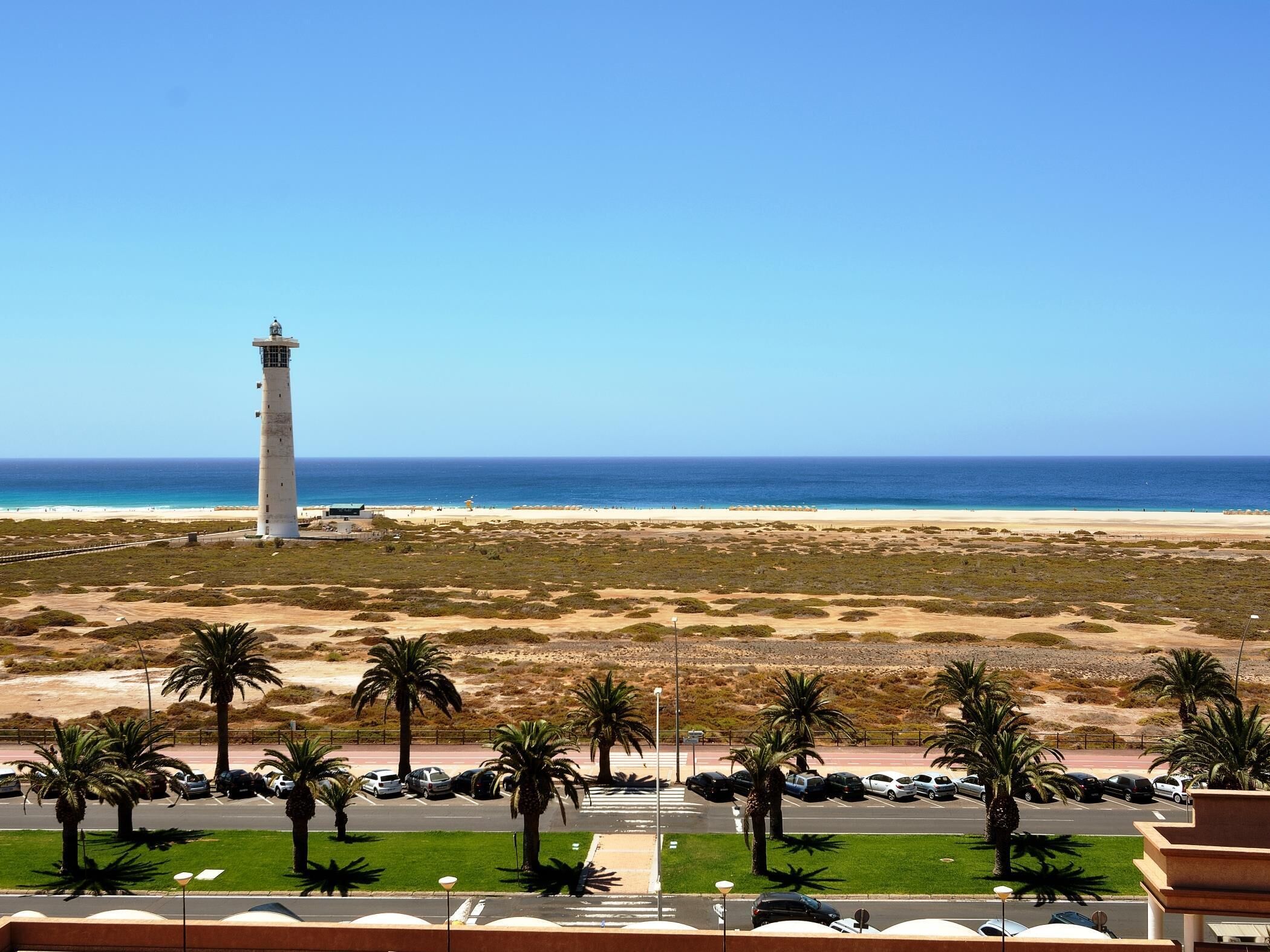 A coastal view with a lighthouse, beach, and parking area with palm trees.