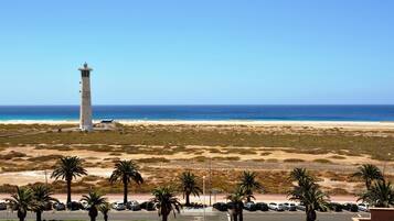 A coastal view with a lighthouse, beach, and parking area with palm trees.