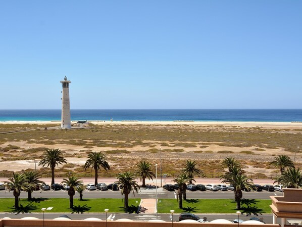 A coastal view with a lighthouse, beach, palm trees, and a parking area.