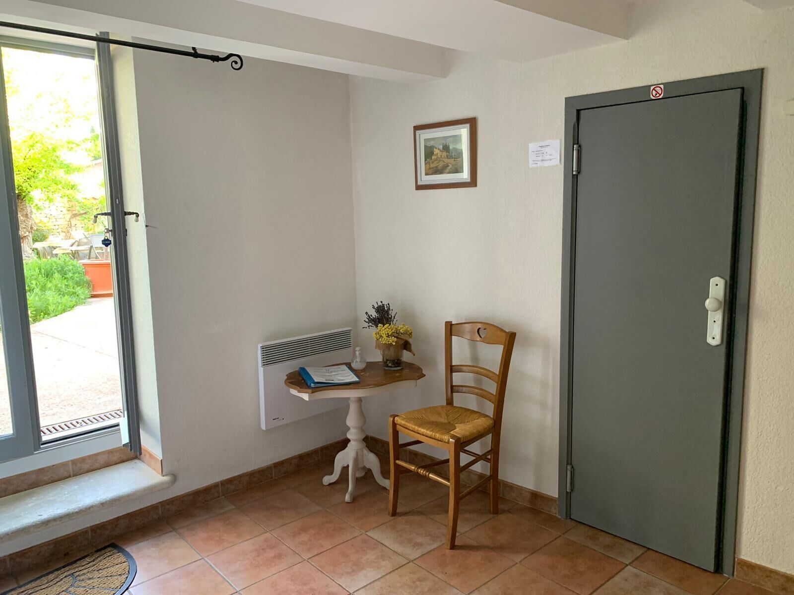 A small room with a window, a door, a radiator, a chair, a small table with a book and flowers, and a framed picture on the wall.
