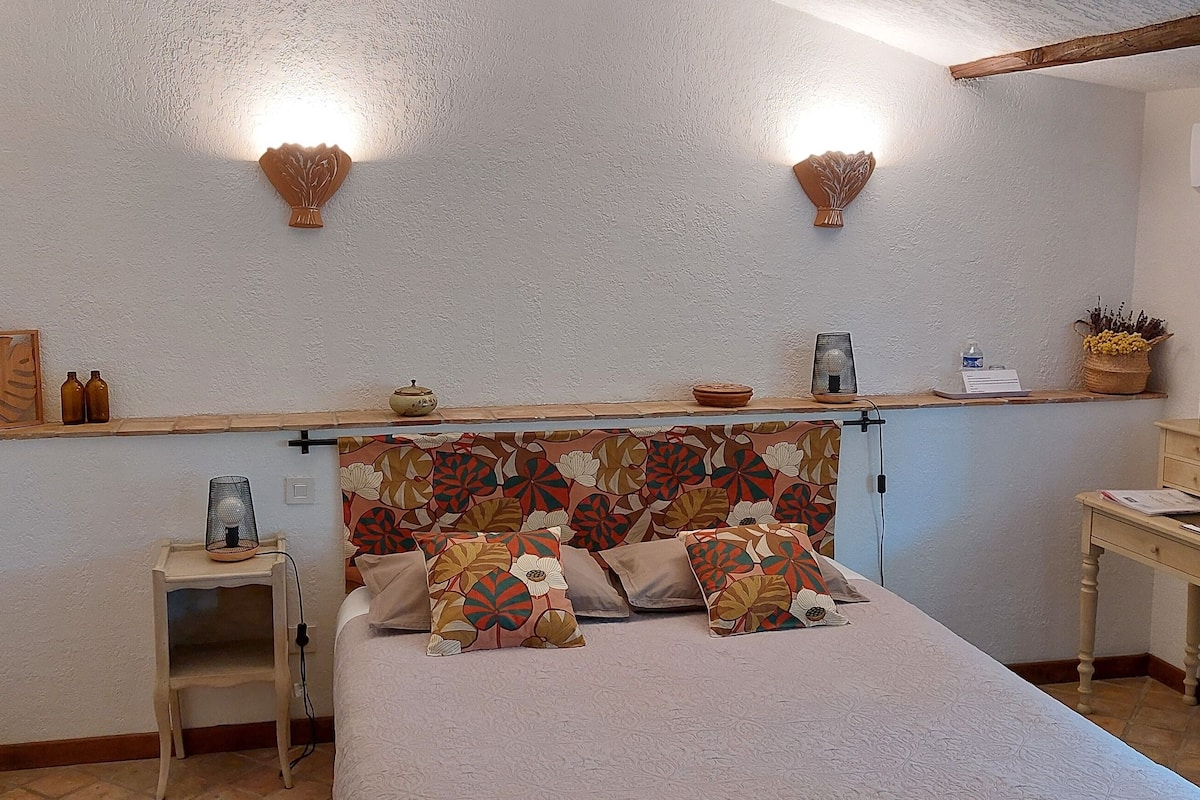 A bedroom with a bed, bedside table, lamp, wall-mounted lights, and a small shelf with decorative items.