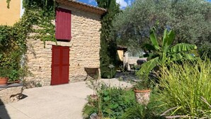 A stone building with a red door and a red shutter, surrounded by potted plants and greenery. - Bastide Sainte Agnes (Carpentras)