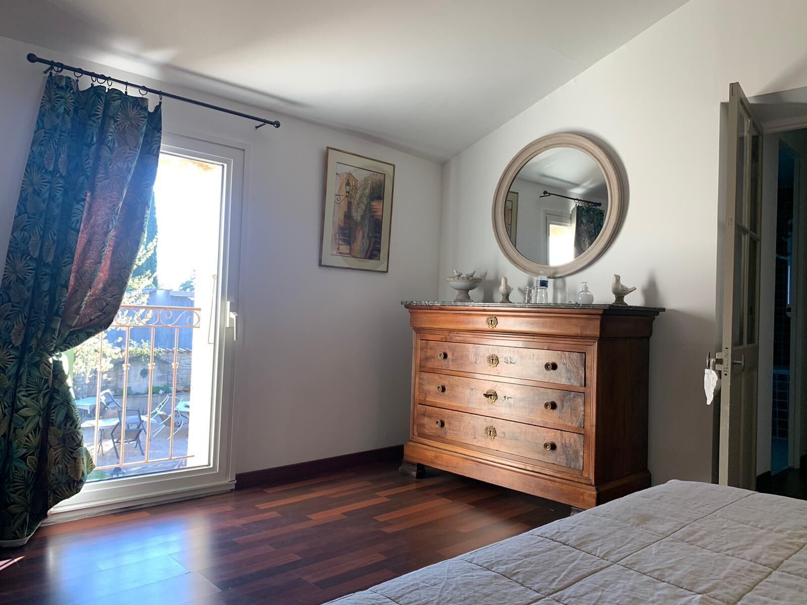 Un dormitorio con tocador de madera, una cama, un ventanal con cortinas, un cuadro en la pared y un espejo.