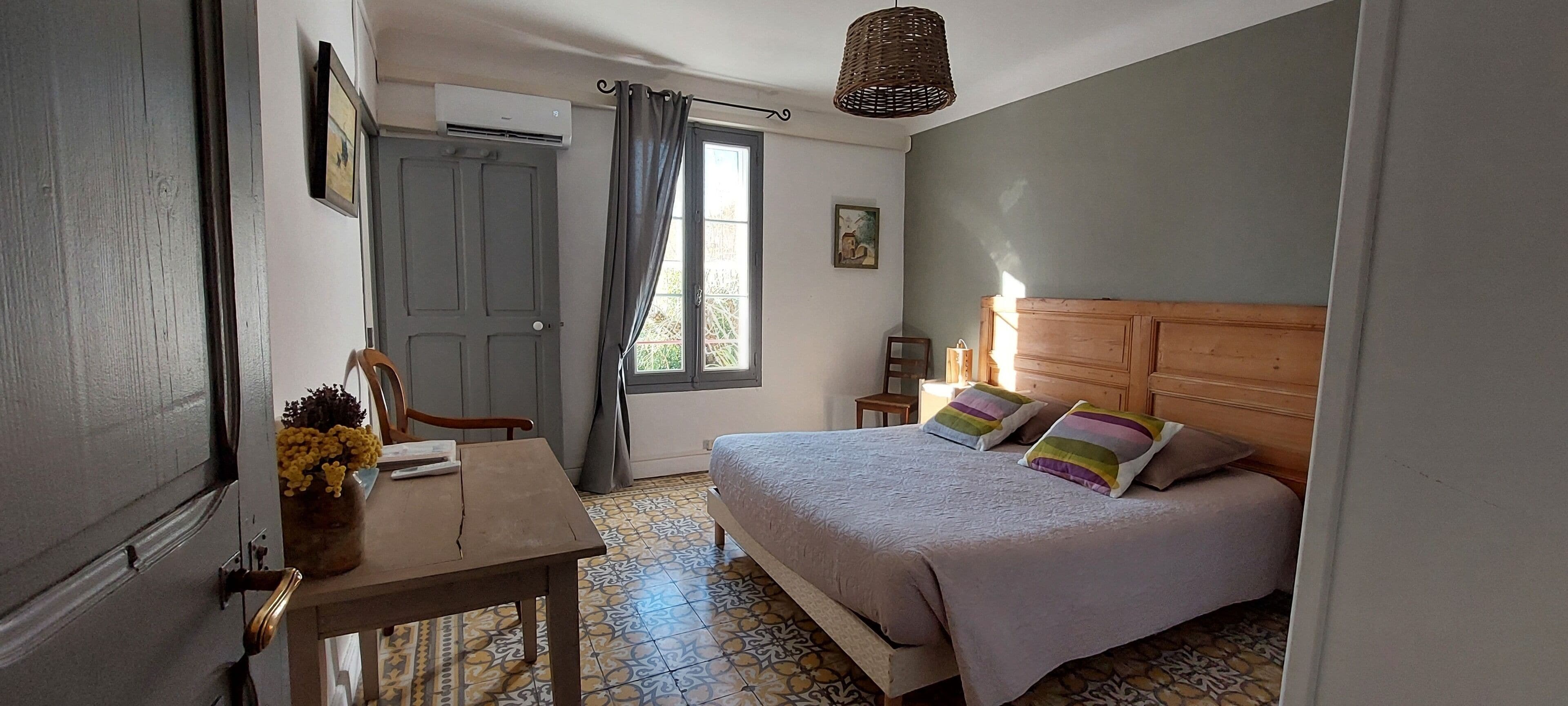 A bedroom with a bed, a wooden headboard, a small table with a vase of flowers, a chair, and a window with curtains.