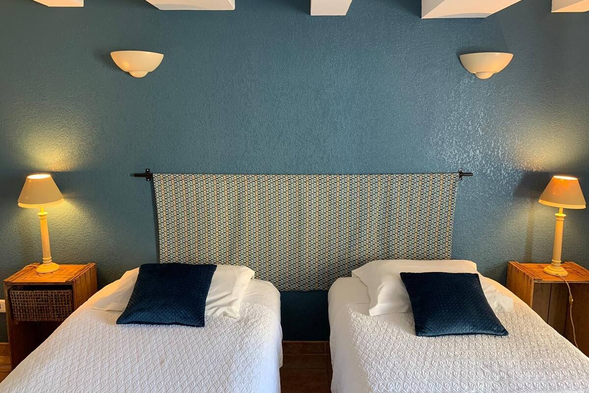 A hotel room with two single beds, each with a dark pillow and a white textured bedspread. There are two bedside lamps on wooden nightstands, and a patterned curtain hangs between the beds.