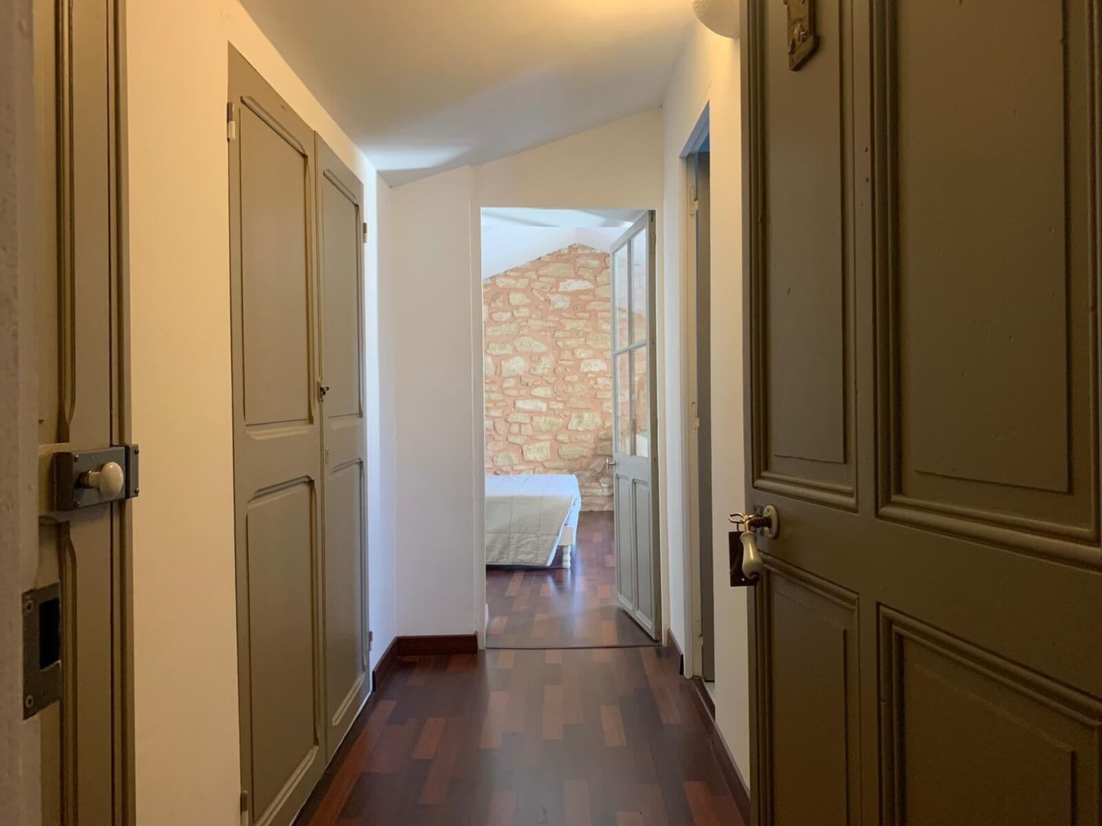 A hallway with wooden floors and doors leading to a room with a stone wall and a bed.