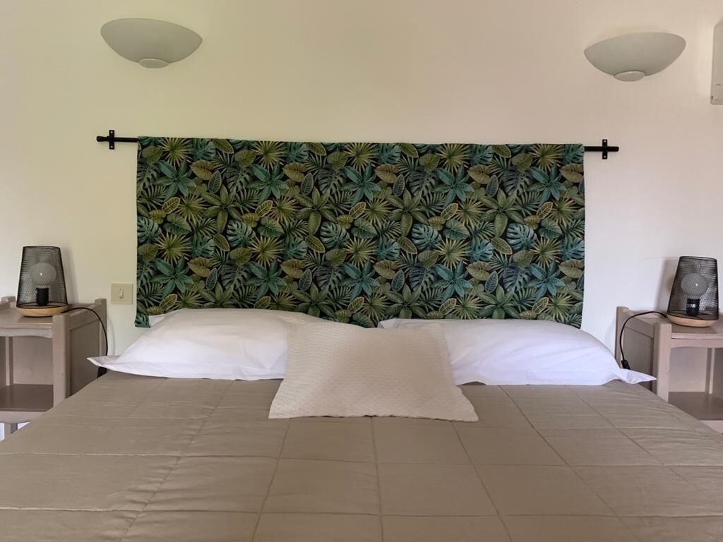 Een bed met hoofdeinde met een tropisch bladmotief, twee nachtkastjes met lampen en een neutraal gekleurd beddegoed.