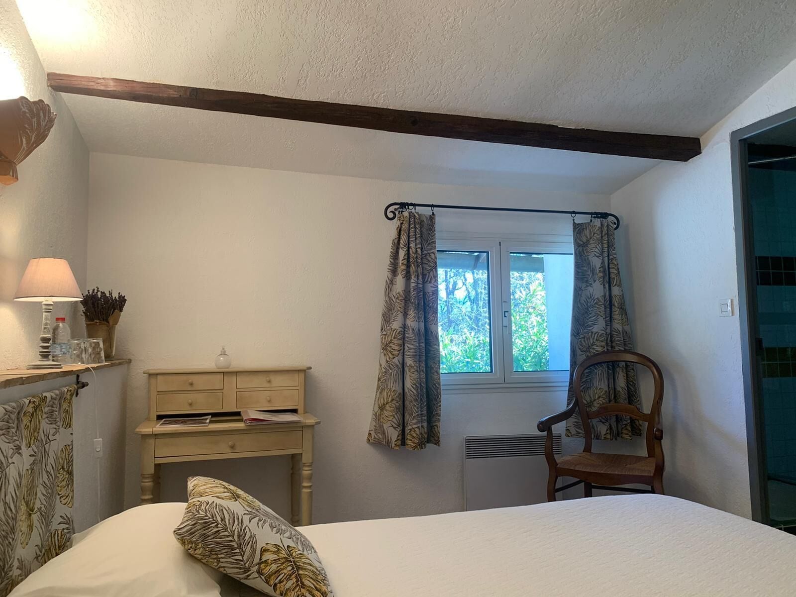 Une chambre à coucher avec un lit, une chaise en bois, un bureau avec une lampe et une fenêtre avec des rideaux.