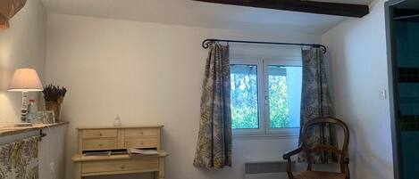 A bedroom with a bed, a wooden chair, a desk with a lamp, and a window with curtains.