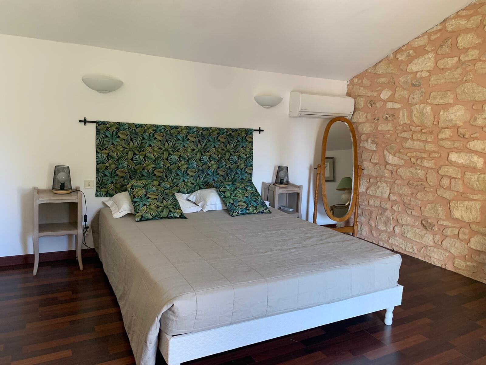 A bedroom with a bed, a nightstand, a mirror, and a wall with a stone pattern.