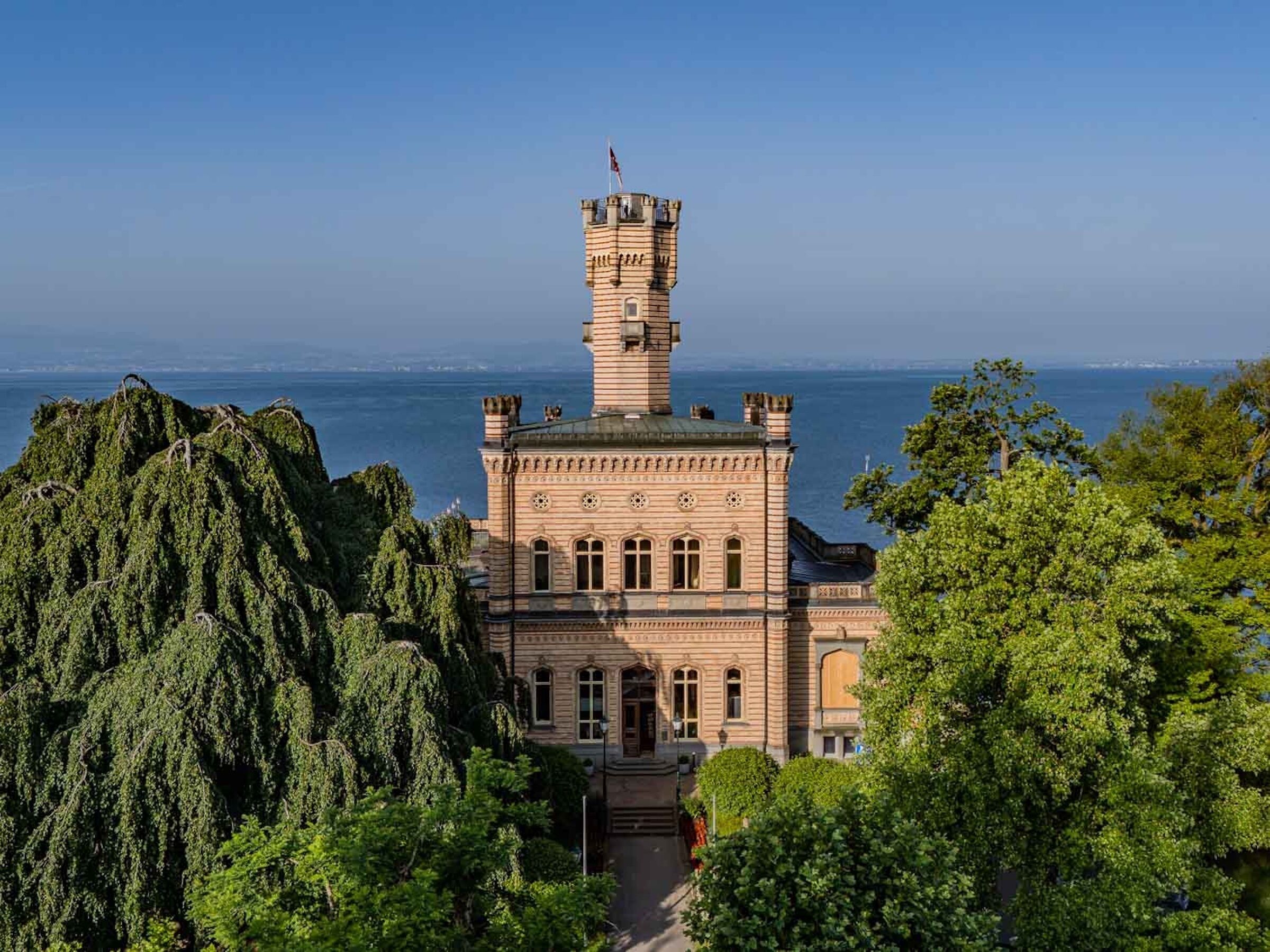 A historic building with a prominent tower, surrounded by lush greenery and overlooking a body of water.
