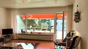 Ein modernes Wohnzimmer mit Fernseher, Glastisch, Sessel, Heizkörper und einem Balkon mit Blick auf Grünflächen.