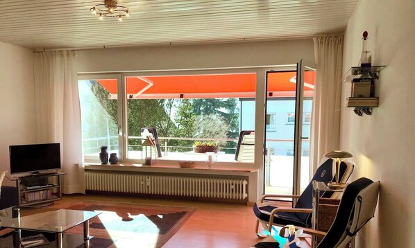 Un salon moderne avec un téléviseur, une table basse en verre, un fauteuil, un radiateur et un balcon donnant sur un espace verdoyant.