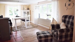 Un salon chaleureux avec un fauteuil à carreaux, une table à manger avec des chaises et une grande fenêtre avec des rideaux.
