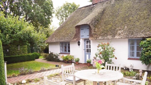 Une petite maison au toit de chaume, avec un jardin, une table ronde en bois et des chaises blanches.