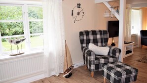 Un salon avec un canapé à carreaux, une petite décoration représentant un voilier, une horloge et une vue sur la verdure à l’extérieur.