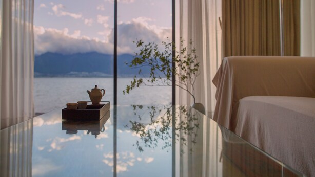 Une chambre d’hôtel avec un lit, une table en verre sur laquelle se trouvent une théière et des tasses, et une vue sur la mer par de grandes fenêtres.