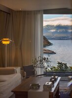 A room with a large window offering a view of a coastal landscape with mountains and a body of water.