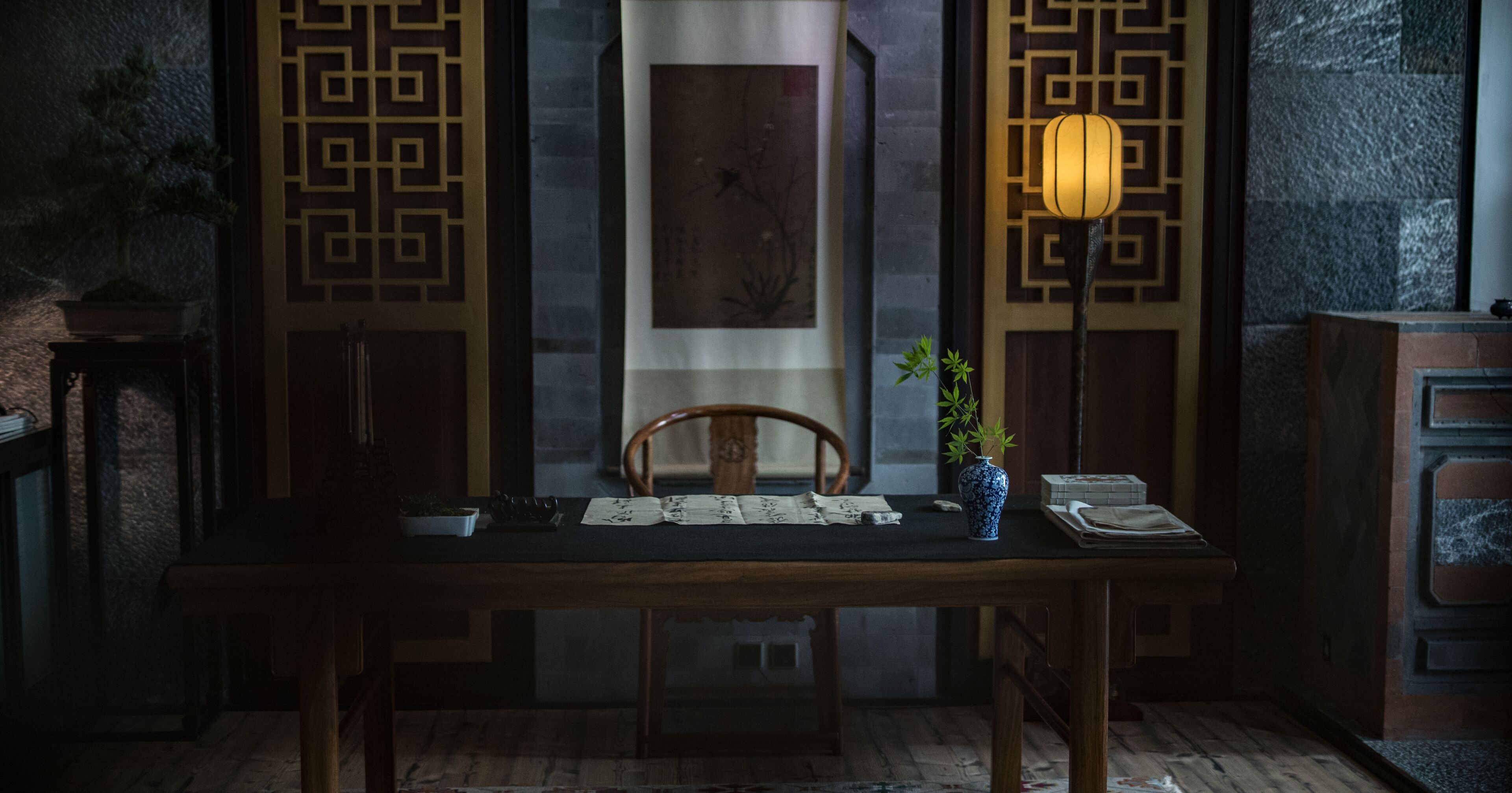 A traditional wooden table with a vase of green plants, a lamp, and a framed picture on the wall behind it.