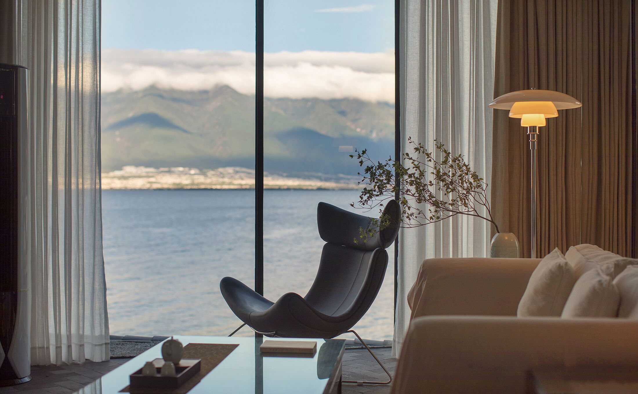Un salón moderno con vistas al mar y a las montañas, una silla negra, un sofá beige y una lámpara de pie.