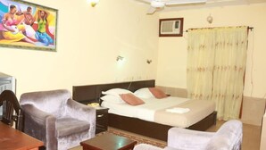 A hotel room with a bed, two armchairs, a wooden table, a ceiling fan, an air conditioning unit, and a painting on the wall.