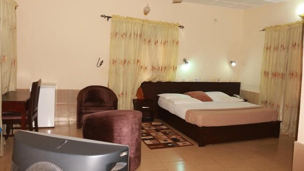 A hotel room with a bed, a desk, a chair, a small table, a fan, and curtains.