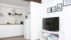 A modern kitchen with white cabinets, a microwave, and a clock on the wall. There is a flat-screen TV on a white stand with a few picture frames above it.