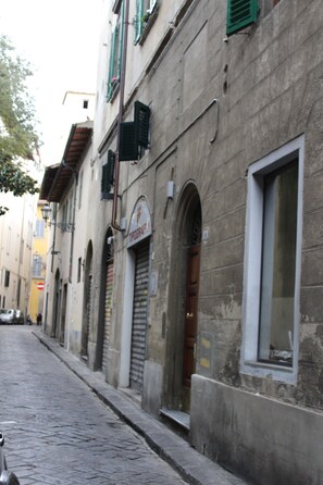 Une ruelle étroite pavée, bordée de bâtiments anciens en pierre, dont certains sont dotés de portes en arc de cercle et de volets verts.