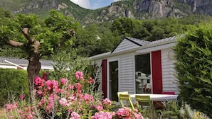 Ein kleines, weißes Gebäude mit roten Fensterläden, umgeben von üppigem Grün und rosa Blüten.