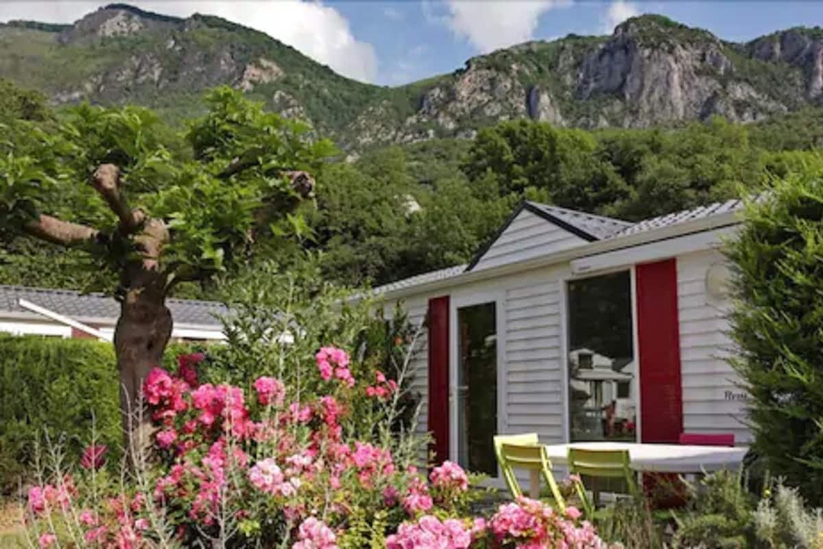 Un pequeño edificio blanco con contraventanas rojas, rodeado de exuberante follaje y flores rosadas.