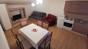 A compact living space with a dining area, a sofa, a red armchair, a television, a small kitchen with a stove, and a ceiling light fixture. - Baka Ljuba (Kutjevo)
