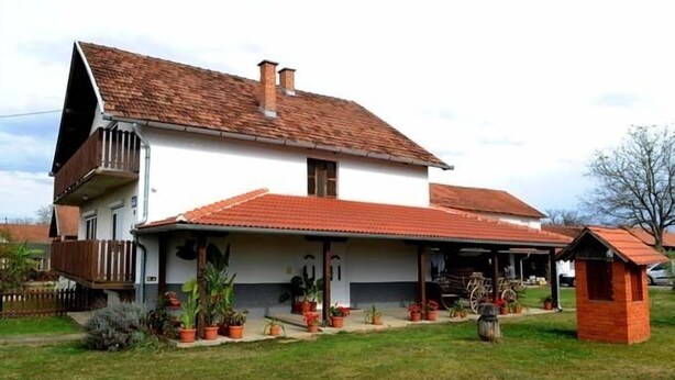 A two-story house with a sloped roof, a covered porch, and a small attached shed. - Baka Ljuba (Kutjevo)